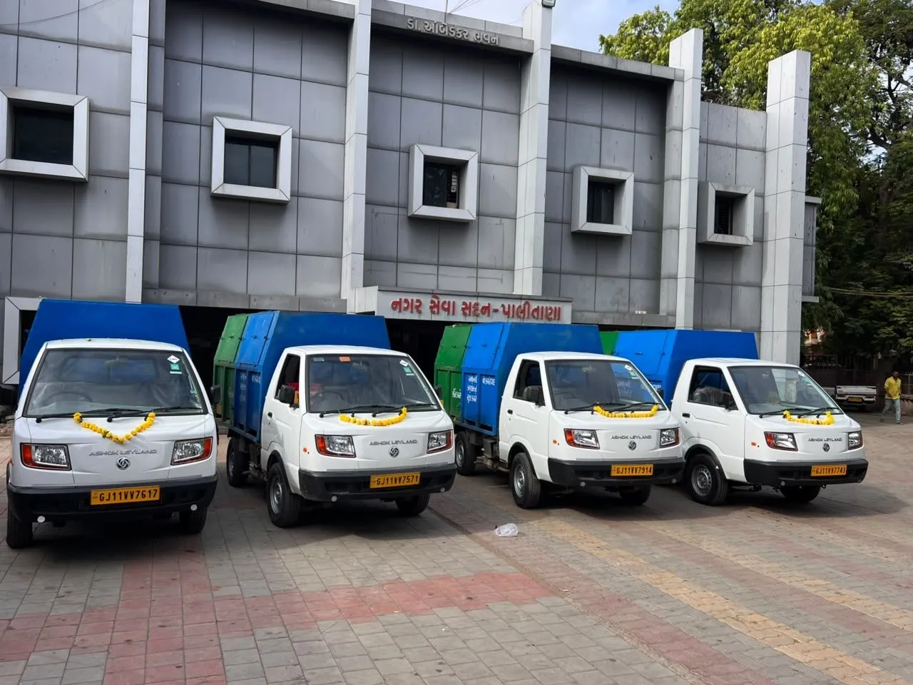 Newly inducted door-to-door collection vehicles, garlanded with marigolds, parked in a row outside a nagarpalika building