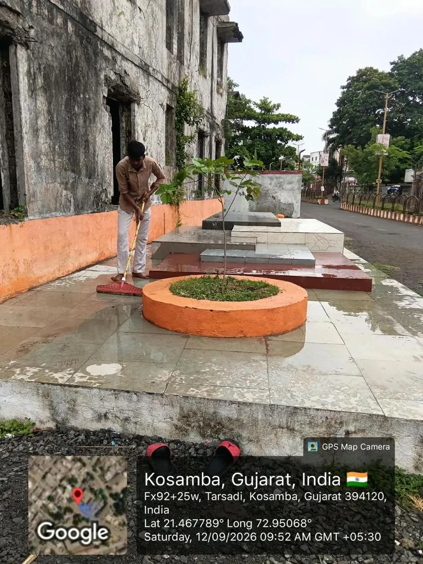 A sanitation worker cleaning a paved public plaza beside a roadside memorial at Kosamba